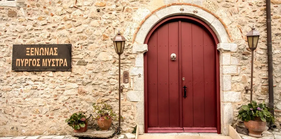 Η κόκκινη τοξωτή πόρτα στον πέτρινο τοίχο με την ταμπέλα του ξενώνα και δύο φανάρια